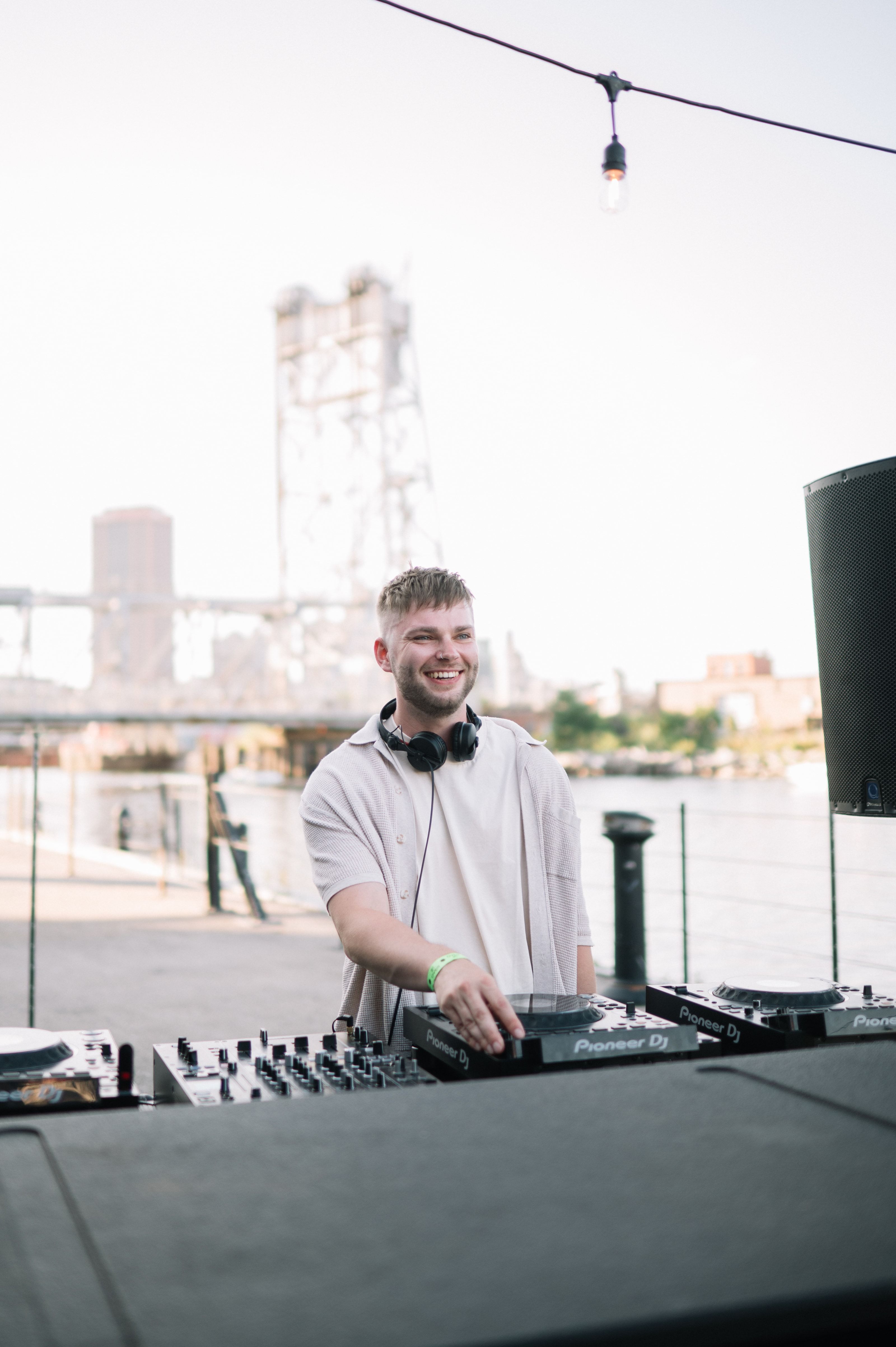 JohnLuc smiling behind his DJ equipment outdoors, with Buffalo’s grain elevators behind him.