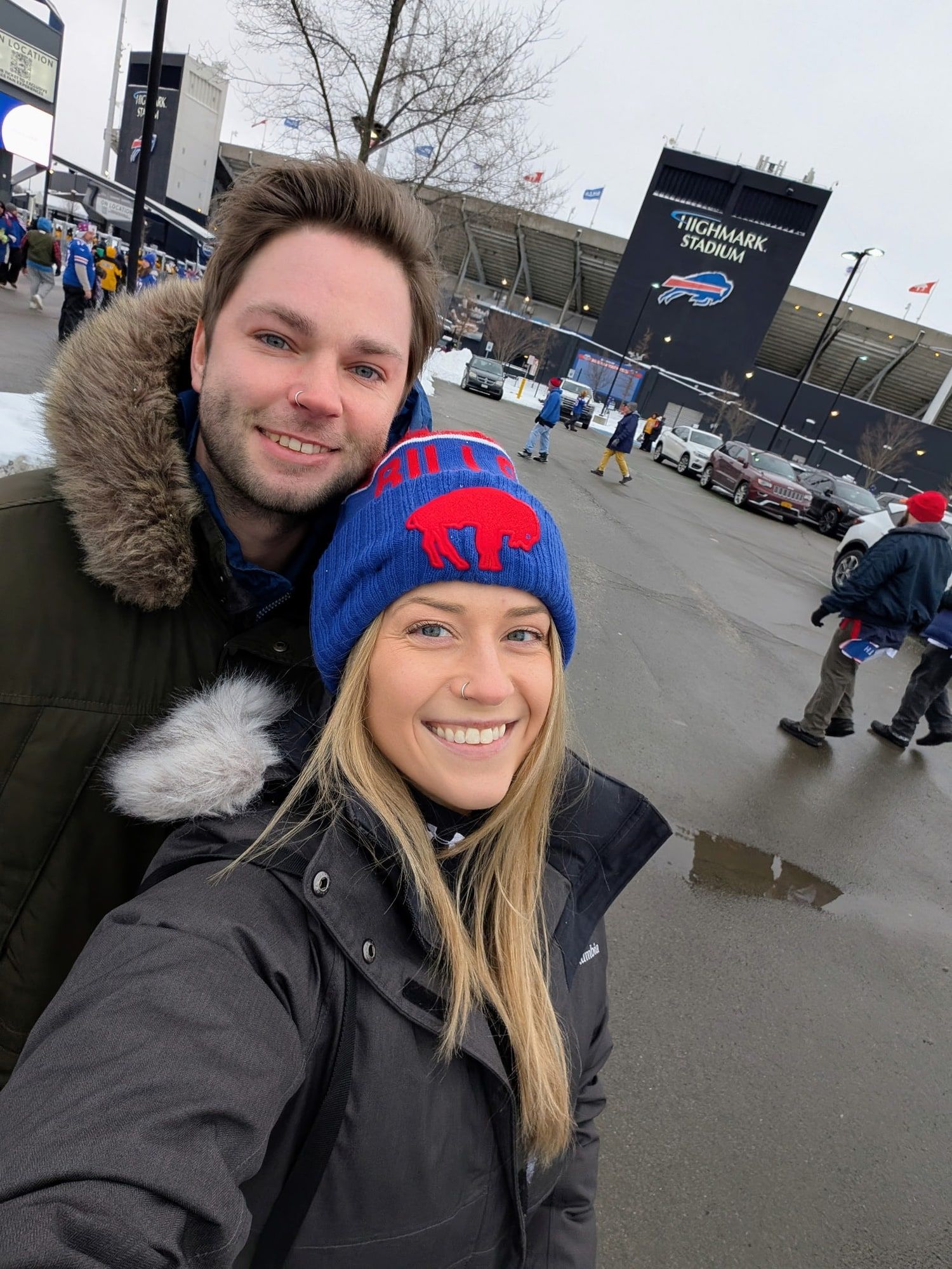 JohnLuc and Katelyn wearing winter coats and Buffalo Bills gear outside Highmark Stadium.